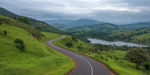 Fototapeta premium A winding road with a lake in the background. The road is surrounded by lush green hills and trees. The scene has a peaceful and serene atmosphere