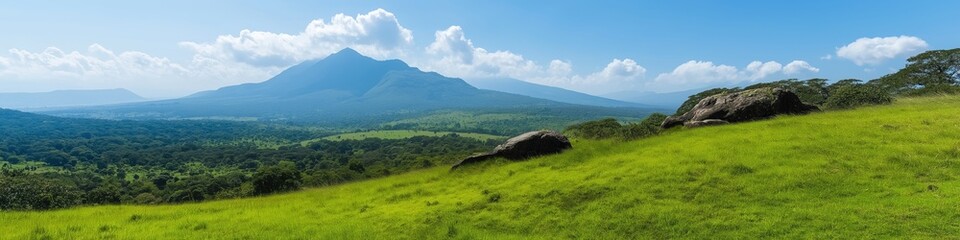 Obraz premium A mountain range is visible in the distance with a large rock on the hillside. The sky is clear and blue, and the grass is lush and green. Concept of tranquility and natural beauty