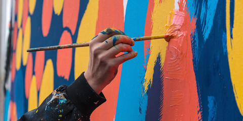 Naklejka premium Young Artist Creating Vibrant Street Mural. Close-up of an artist's hand applying a stroke of bright magenta paint to a colorful, abstract mural.