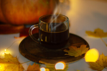 A warm cup of coffee with steam rising, placed on a wooden coaster surrounded by autumn leaves, a pumpkin, and soft glowing lights. A cozy fall atmosphere, perfect for seasonal imagery.