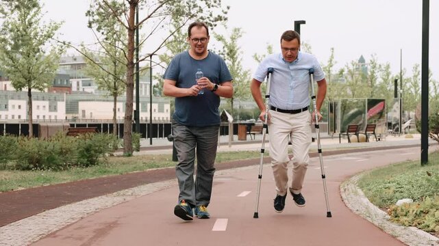 A man barely hobbles on one leg with crutches walking with friend in park. Adult caucasian male walking slowly down the street, speaking with partner. Disabled person relationship concept