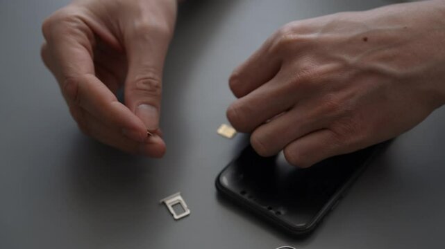 Close-up top view of unrecognizable man carefully swapping sim card inside smartphone, sitting at table with grey surface with extraction tray nearby during mobile service transition, slow motion.