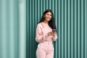 Happy Asian teen girl wearing pink outfit using mobile phone apps outdoors looking at camera....
