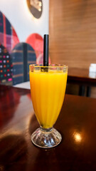 A vibrant glass of fresh mango juice with a straw, placed on a polished wooden table, against a colorful blurred background
