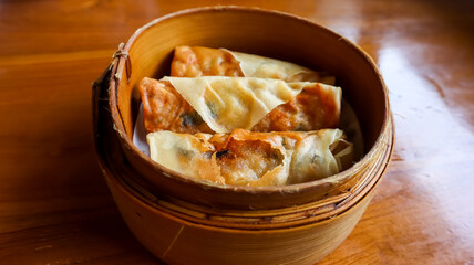 Three spring rolls or lumpia in a bamboo steamer basket, placed on a wooden table