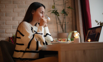 A woman in a cozy sweater enjoys a warm drink while sitting at a table with a laptop, salad, and modern home decor. The atmosphere is relaxed and comfortable, ideal for work or leisure moments.