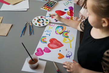 Two children are involved in an artistic activity, painting colorful hearts, rainbows, and the word 'LOVE' on a sheet of paper, using brushes, paint palettes, and vibrant watercolors.