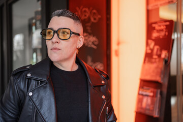 Gender-fluid person wearing yellow sunglasses and black leather jacket posing near red neon sign