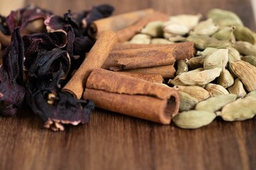 A close-up of vibrant spices: dried hibiscus, cinnamon sticks, and green cardamom pods on a wooden surface, showcasing their rich textures and colors.