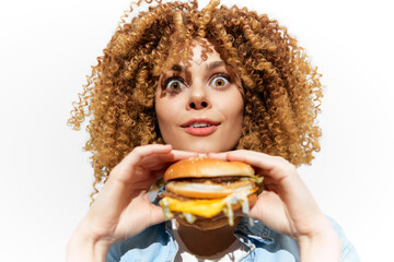 Naklejka premium Young woman with curly hair excitedly holds a delicious cheeseburger, showcasing fresh ingredients and vibrant colors, on a white background
