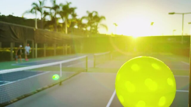 Close-up shot of pickleball ball, people playing pickleball during sunset