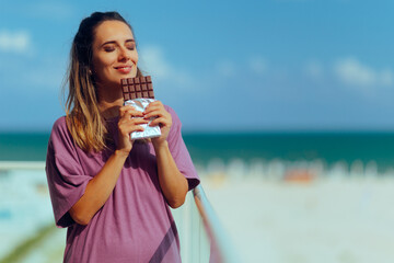Woman Eating a Chocolate Bar in the Balcony. Lady craving a sweet dessert treat at her beach home
