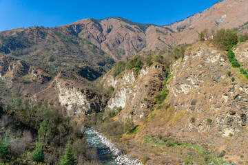 Obraz premium winding, rocky river flows through a deep, arid mountain valley, flanked by steep, eroded cliffs & patches of sparse green & brown, dry vegetation. bright blue sky, harsh beauty of the high mountains.