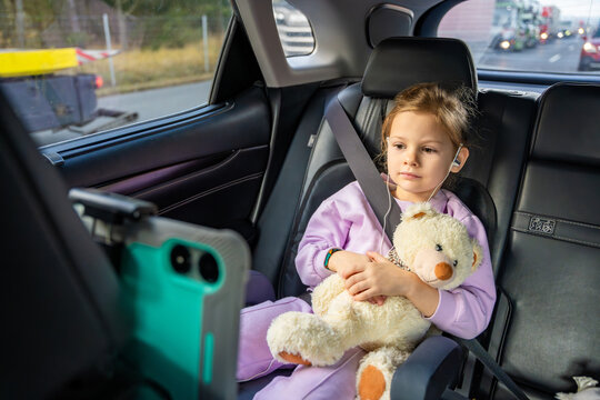 Little girl with teddy bear, tablet and headphones in the car during travel on vacation. Entertainment in the transport for child