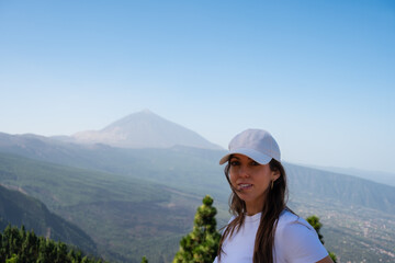 A woman is standing on a mountain top with a hat on. The sky is clear and the sun is shining