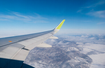 Fototapeta premium View from an airplane window, with the wing in the foreground and a landscape of snowy mountains and clouds in the background.