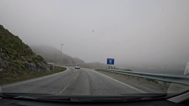 Driving through the tundra in northern Norway on E69 road. The sea coastline is on the right, while grassy hills and rocky cliffs rise on the left. Mist and fog add a mysterious atmosphere.