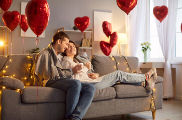 Portrait of young cheerful man and woman hugging indoors sitting on sofa in living room at home with a cup of beverage. Happy couple in love. Relationships, romance and Valentines day concept.