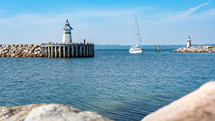 Blick auf die Hafeneinfahrt und die Signallichter von Ebeltoft während gerade ein weißes Segelboot einläuft, Steine unscharf im Vordergrund, horizontal