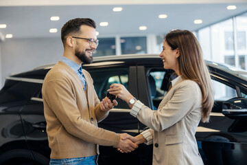 Side view of smiling saleswoman giving car keys to customer and shaking hands with him at car salon.