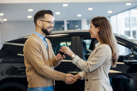 Side view of smiling saleswoman giving car keys to customer and shaking hands with him at car salon. - Powered by Adobe