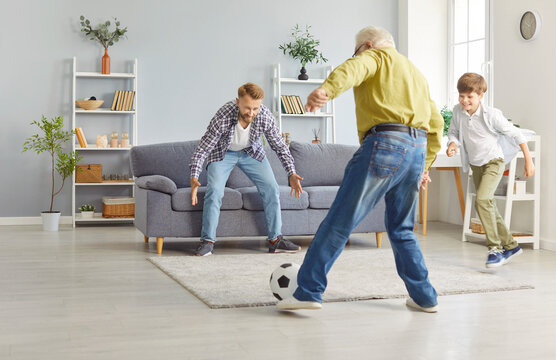 Happy family playing football. In bright living room, three generations of men are having fun playing and passing ball. Lively and nimble grandfather kicks ball towards his son and little grandson.