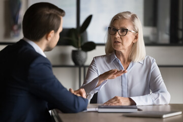 Confident mature business professional woman talking to younger male colleague, explaining work...