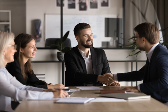 Two smiling millennial businessmen shaking hands, close deal, make agreement after successful negotiations in office boardroom with colleagues. Handshake gesture of commitment, partnership and trust