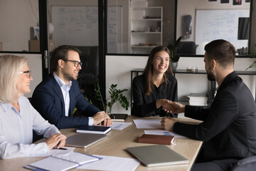 Two business partners shaking hands in conference room at table during group meeting with project...