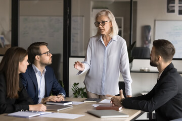 Mature 55s company representative or coach leading negotiations or training for staff, standing in front of partners, makes speech, explain task details, project conditions gathered in conference room
