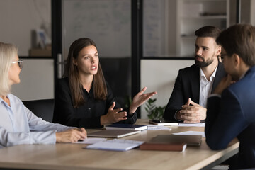 Serious young and mature colleagues brainstorm at conference table, listening speech of female team...