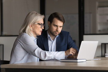 Two 35s and 55s teammates sitting at desk, looking at computer monitor, working on common task, collaborate on joint project, discuss online marketing strategy met in conference room of modern office
