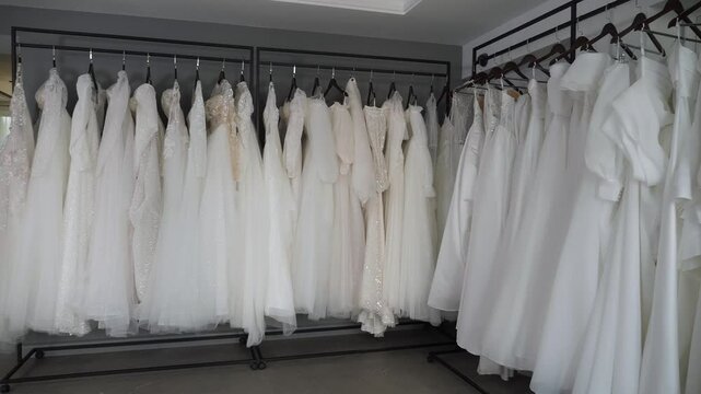 Many different wedding dresses are hanging on hangers in a modern wedding salon. Various wedding dresses. Wedding fashion. Stable movement of the camera backwards. Slow motion.