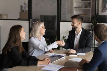 Older and younger businesspeople, teammates shaking hands seated at desk in office take part in...