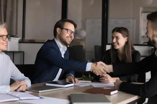 Smiling male colleagues, shareholders shake hands close deal at meeting with diverse employees in modern office. Businessmen handshake, get acquainted, greeting each other, making business agreement