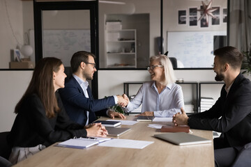 Two business partners, leaders shaking hands seated around office table, start group meeting in...