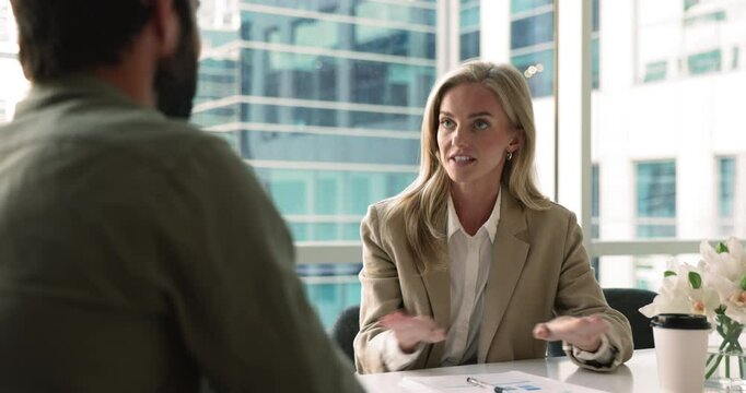 Happy confident blonde business leader woman talking to partner, job candidate, colleague man, speaking with hands moving, sitting at office meeting table, shaking hands with male coworker