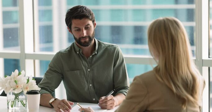 Couple of colleagues talking at workplace table, discussing project marketing paper report. Female manager presenting strategy to male colleague, boss, pointing at document, speaking