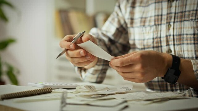 A person sorts through various receipts and documents on a cluttered desk, using a calculator and a pen. Plants are visible in the background, creating a cozy workspace atmosphere