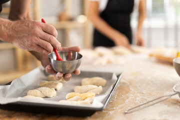 Close up of baker hands brushing uncooked dough pieces on baking sheet with oil before baking in oven