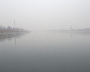 Naklejka premium serene lake shrouded in fog, reflecting muted colors of industrial zone nearby. atmosphere evokes sense of calm amidst haze