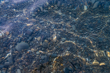 Underwater pebble backgrounds. Colorful stones sea bottom in the sun rays. View from above through clear water. Seascape wallpaper texture. Defocus.