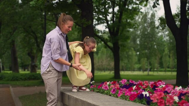 Woman helps young girl with Down syndrome lean over stone wall to smell vibrant flowers in park. Green trees and natural background enhance peaceful atmosphere. Concept of social inclusion and