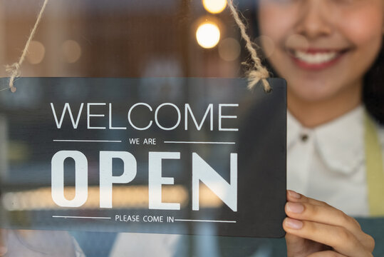 beautiful female business retail owner in medical face mask hanging open wooden sign board at the entrance door of the shop and ready to service customer.
