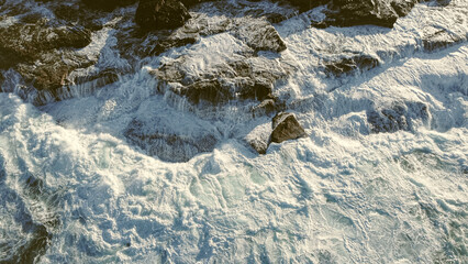An aerial view of powerful ocean waves crashing against rugged rocks, creating dramatic splashes and foamy textures. The sunlight highlights the textures, evoking a sense of raw natural power and beau