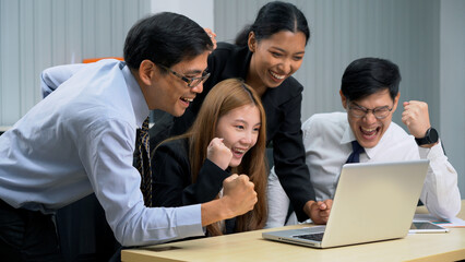 Fototapeta premium Group of happy asian business people sitting on a table raised fists cheering at laptop celebrating achievement goals to success in meeting at office