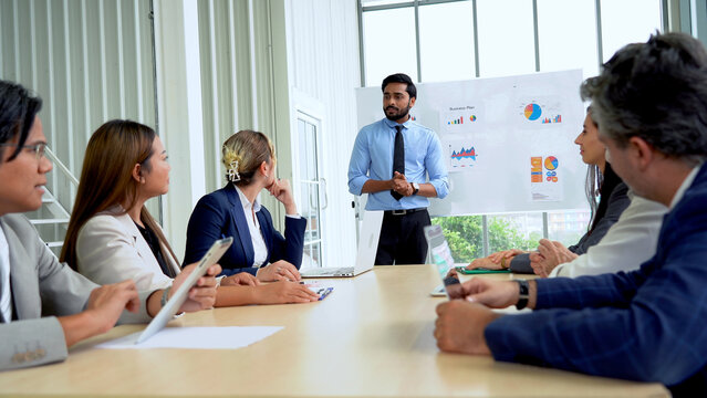 Young indian business manager standing and presenting to multiracial group of diversity colleagues in front of white board in meeting at boardroom in office . leader