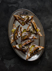 Delicious appetizer, tapas, snack - sandwiches with rye bread, canned sardines, gherkins and cream cheese on a dark background, top view