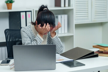 Asian office worker overwhelmed with work stress, holding head in hands at desk with laptop,...