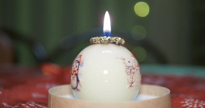 Holiday season. A decorative candle in the form of a ball with painted Christmas pictures stands on a table against a background of flickering multicolored lights. Hand held.